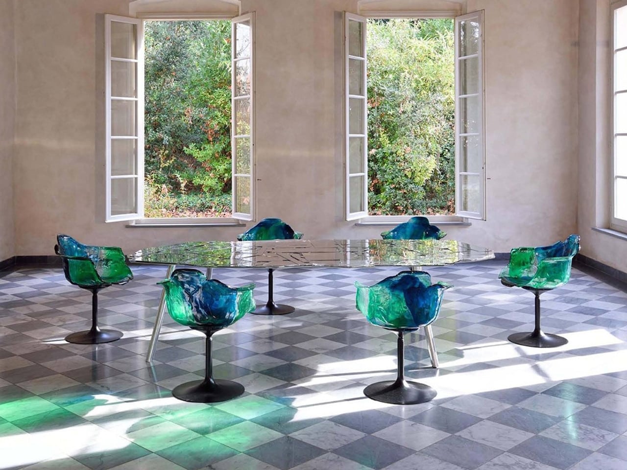 Een set Ella stoelen in groen en blauw polycarbonaat staat rond een glanzende tafel in een ruime, lichte eetkamer met een klassiek patroon op de vloer. Het transparante materiaal van de stoelen creëert een spel van kleuren en licht, dat samen met de open ramen een verbinding met de natuur benadrukt. Dit ontwerp van Edra voegt een eigentijdse en kunstzinnige uitstraling toe aan het interieur.