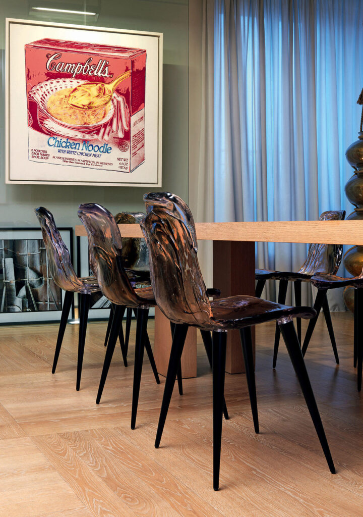 Deze afbeelding toont de Gilda B. Stoelen van Edra in een moderne eetkamer, gecombineerd met een houten tafel en kunstwerken aan de muur. De transparante amberkleurige stoelen vormen een speels contrast met het kunstwerk van Campbell's soep en de minimalistische inrichting van de ruimte. De artistieke vorm en materialen van de stoelen benadrukken hoe ze een verfijnde en creatieve touch toevoegen aan moderne interieurs, perfect voor liefhebbers van design en kunst.