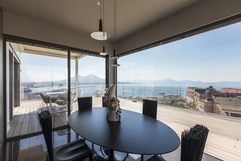 Een moderne eetkamer met een ovale houten tafel en zwarte en witte Jenette-stoelen van Edra. De ruimte wordt gekenmerkt door gedetailleerd plafondwerk, grote ramen en een speels kunstwerk, wat een balans creëert tussen elegantie en creativiteit.