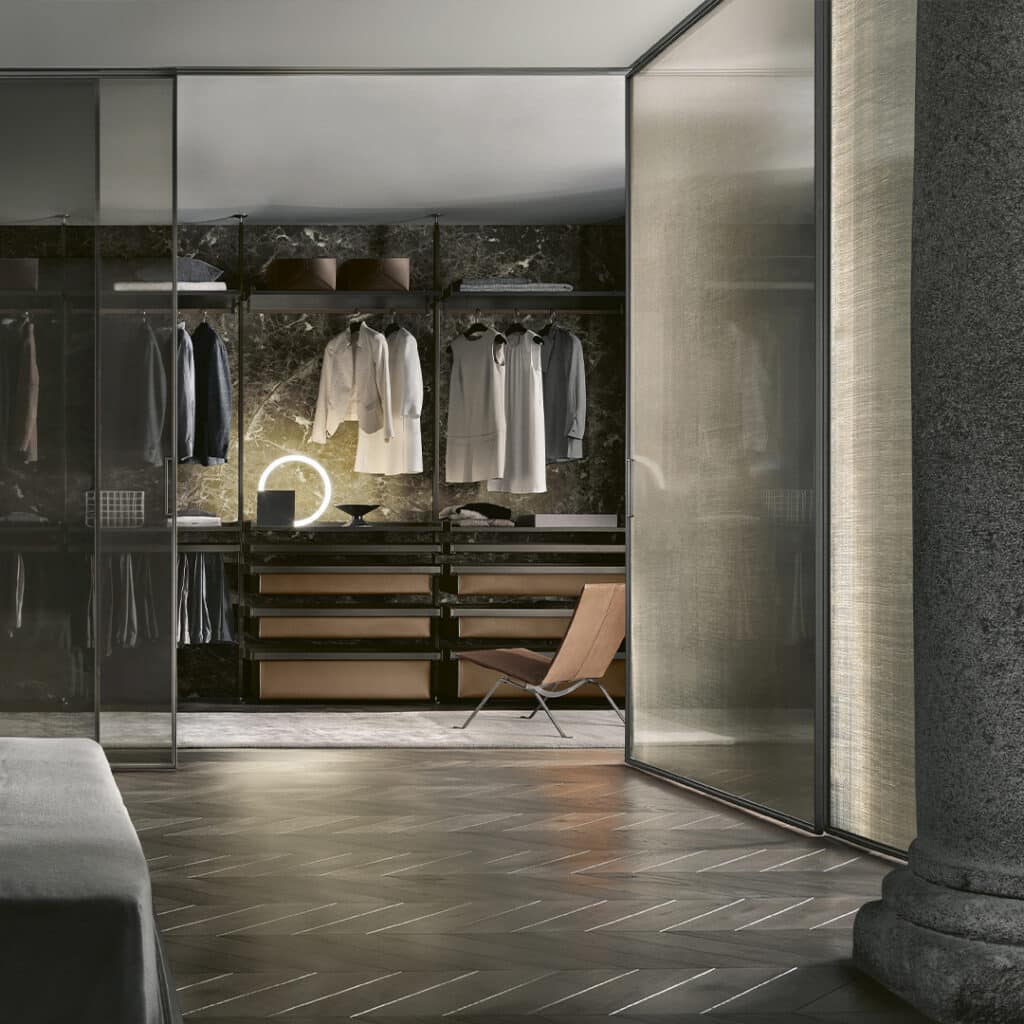 enit Walk-in Closet van Rimadesio, compleet met glazen deuren en een verfijnde, minimalistische indeling. De kast heeft een elegante, moderne uitstraling met op maat gemaakte planken en lades, perfect voor het opbergen van kleding en accessoires. De ruimte wordt verlicht door subtiele verlichting, wat een warme en luxe sfeer creëert. De kast is zorgvuldig ontworpen met premium materialen zoals glas en metaal, en biedt zowel functionaliteit als esthetiek in een exclusief interieur. De minimalistische stoel en het marmeren achterpaneel voegen extra verfijning toe aan de ruimte, die zowel praktisch als visueel indrukwekkend is.