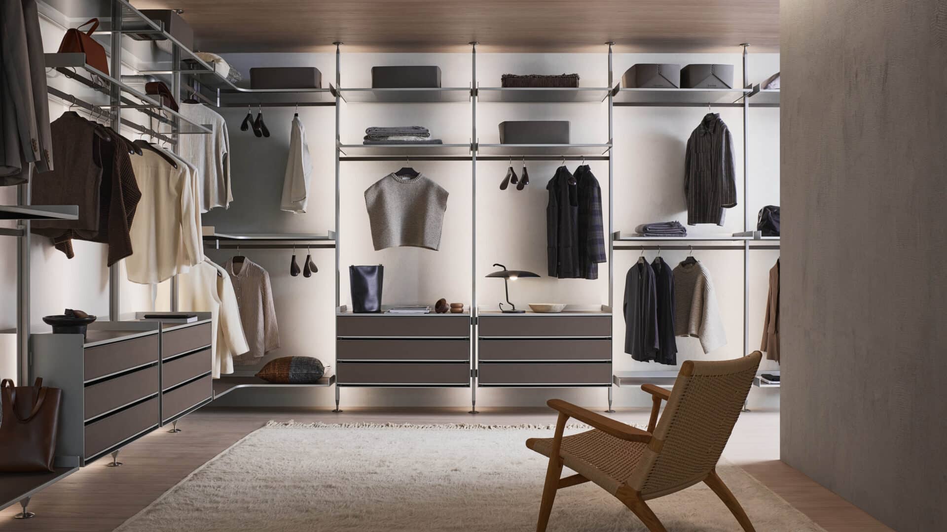 modulaire Zenit Walk-in Closet van Rimadesio, die een luxe en minimalistisch ontwerp biedt. De kast is uitgerust met glanzende ladefronten, stijlvolle planken en opbergruimte voor kleding, accessoires en schoenen. De zachte verlichting benadrukt de strakke lijnen en het open ontwerp van het systeem. De ruimte is zorgvuldig georganiseerd, met voldoende opbergruimte voor een gestructureerde en elegante indeling. Het verfijnde, moderne design wordt verder versterkt door de eenvoudige stoel en luxueuze materialen zoals het hout en metaal, die samen een elegante sfeer creëren.